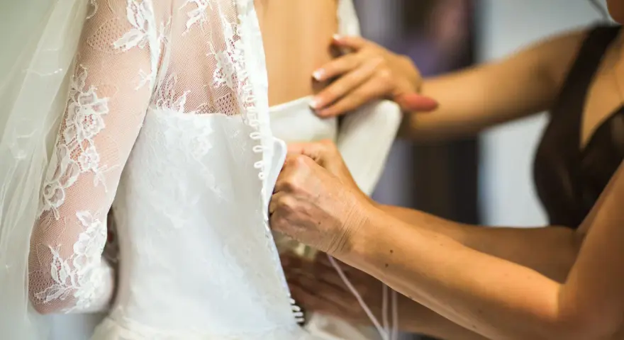 Dettaglio di abiti da sposa riutilizzabili: mani che chiudono bottoncini su un vestito bianco con pizzo e velo.