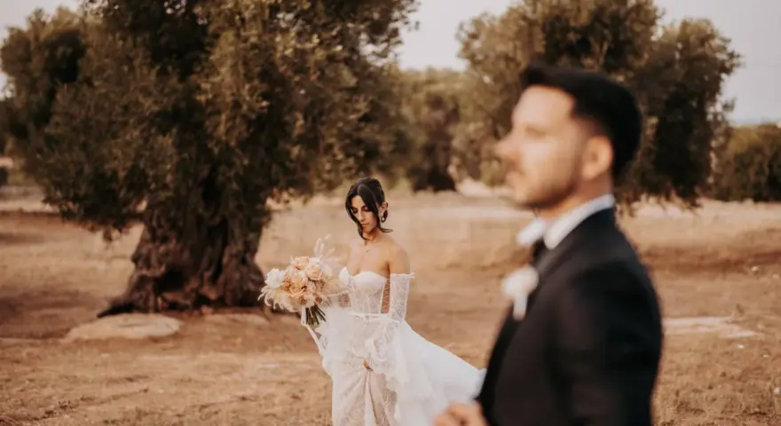 Sposarsi a maggio sotto un ulivo secolare. La sposa con un bouquet di rose pesca, lo sposo in primo piano sfocato.