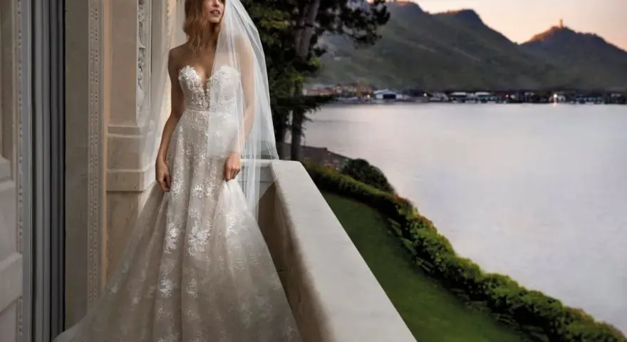 Sposa con abito da sogno, con vari tipi di pizzo che scendono a cascata, su un balcone con vista lago al tramonto.