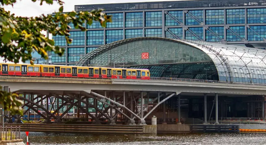 Treno giallo e rosso attraversa un ponte sopra un fiume, con la moderna stazione ferroviaria di Berlino sullo sfondo. Un ottimo modo per capire come muoversi a Berlino.