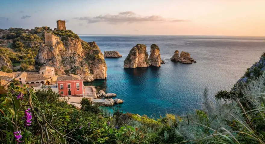 Un borgo con torri antiche e un edificio rosso si affaccia su un mare cristallino, perfetto per le mete estive in Italia.