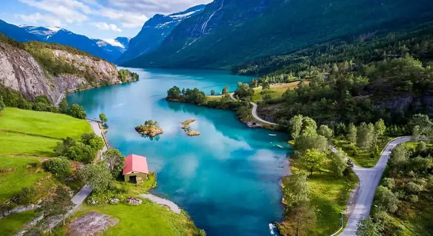 Un lago turchese serpeggia tra montagne verdi e innevate. Un piccolo edificio rosso si affaccia sull'acqua. La crociera fiordi periodo migliore è l'estate per godere di questi panorami.