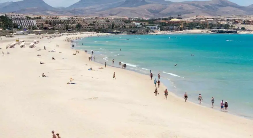 Spiaggia affollata con persone che passeggiano e si rilassano, hotel e montagne sullo sfondo. Questo è il paesaggio di Fuerteventura, dove si trova questo paradiso.