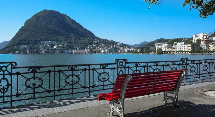 Panoramica del lago di Lugano con Monte San Salvatore sullo sfondo. Una panchina rossa invita a godersi la vista, perfetta per scoprire cosa vedere a Lugano in un giorno.