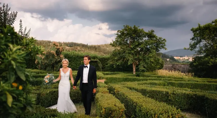 Sposi passeggiano mano nella mano tra i verdi labirinti della **villa di Maiano**, con un bouquet fiorito e uno sfondo di colline toscane.