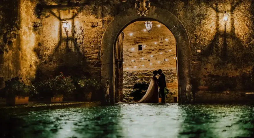 Una coppia di sposi si abbraccia sotto un arco del castello di San Basilio, illuminato da lanterne e luci soffuse. L'acqua riflette l'atmosfera magica.
