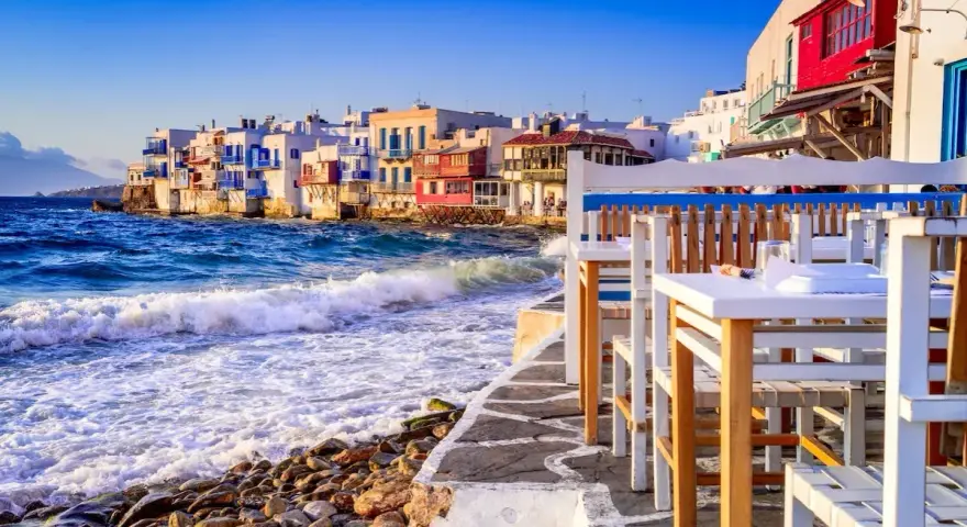 Case vacanza a Mykonos, un'idea per dove andare a vivere all'estero con pochi soldi. Case bianche sul mare, onde che si infrangono sugli scogli, tavoli di un ristorante.