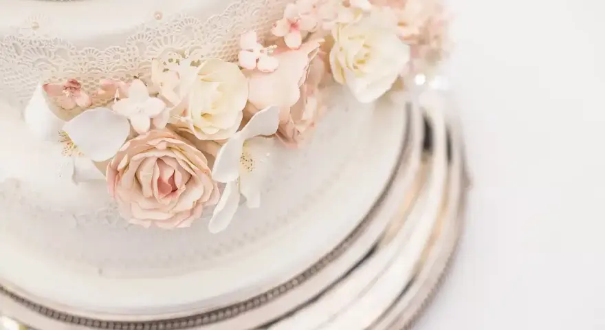 Dettaglio di una torta nuziale a più piani, decorata con pizzo e fiori di zucchero rosa e bianchi, su un elegante vassoio argentato.