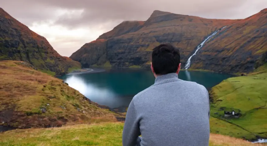 Uomo con maglione grigio ammira un paesaggio mozzafiato delle Isole Faroe, con un lago turchese e una cascata.