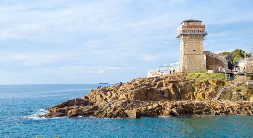 Torre medievale e mare blu a Calafuria, con scogli rocciosi e cielo nuvoloso.