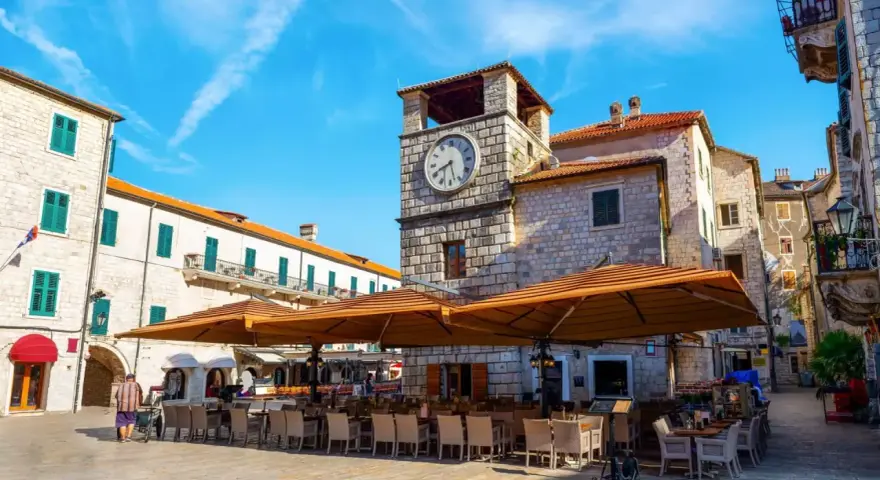 Piazza nel kotor centro storico con torre dell'orologio e tavoli all'aperto di un ristorante.