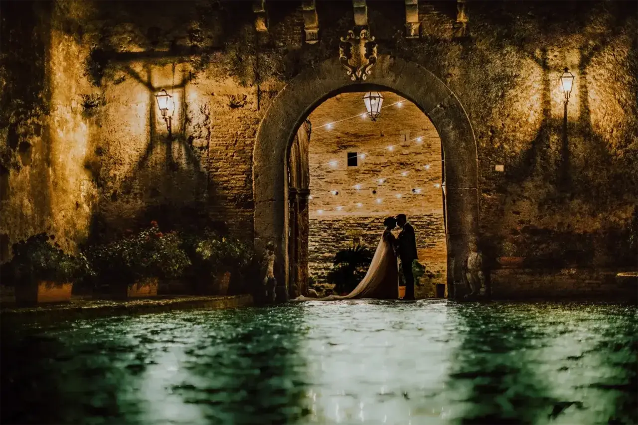 Coppia di sposi abbracciati sotto un arco del castello di San Basilio, con luci soffuse e riflessi sull'acqua.