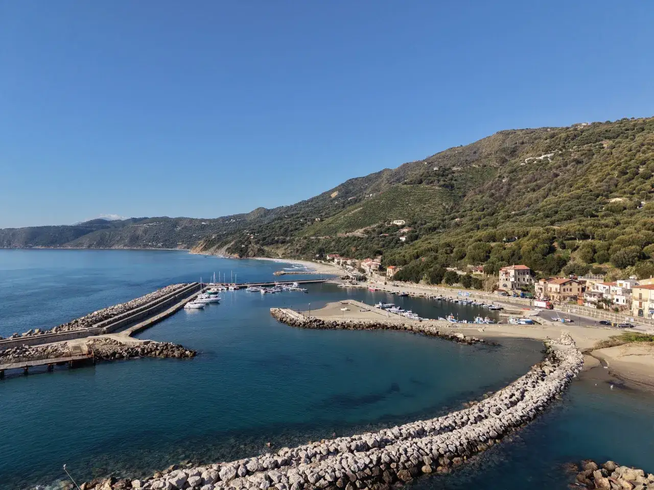 Marina di Pisciotta, con il suo porto turistico, le barche ormeggiate e le case arroccate sulla collina verdeggiante, sotto un cielo azzurro.