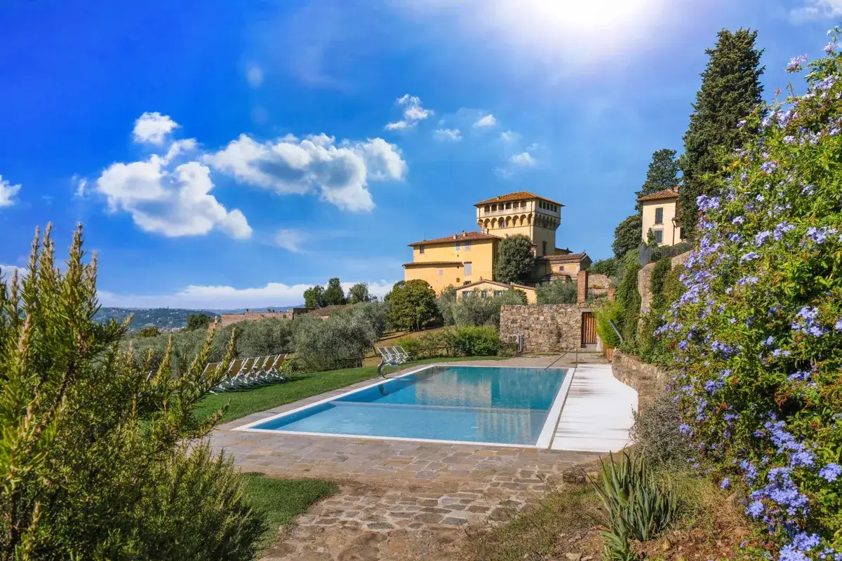 Piscina con vista sulla storica villa di Maiano, circondata da ulivi e fiori blu sotto un cielo azzurro.