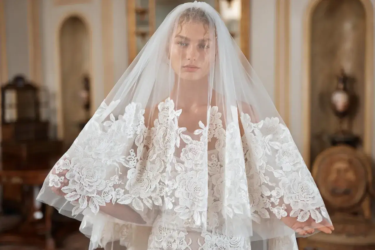 Volto di una sposa con un velo di pizzo ricamato che incornicia il viso. Il velo della sposa, leggero e trasparente, nasconde parzialmente i suoi occhi intensi.