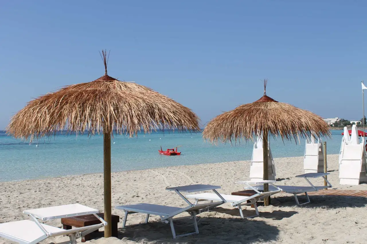 Ombrelloni di paglia e lettini su una spiaggia di sabbia bianca a Punta Prosciutto. Il mare cristallino invita al relax. Scopri i prezzi per goderti questo paradiso.