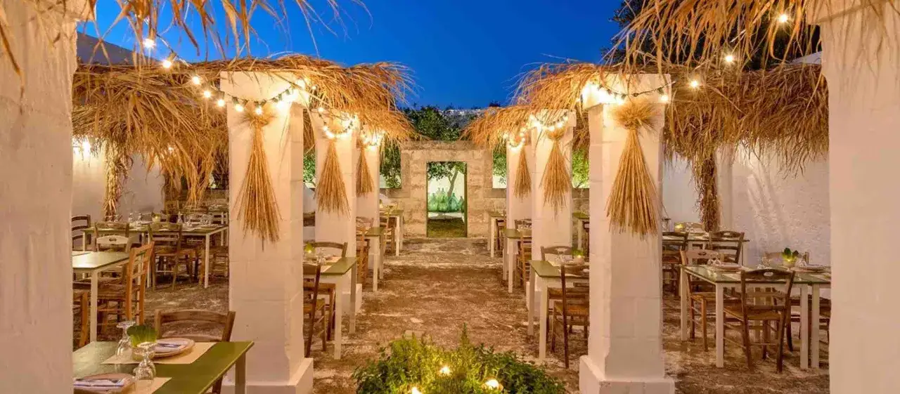 Atmosfera magica in un ristorante stellato Puglia, con tavoli apparecchiati sotto pergolati di paglia illuminati da lucine.