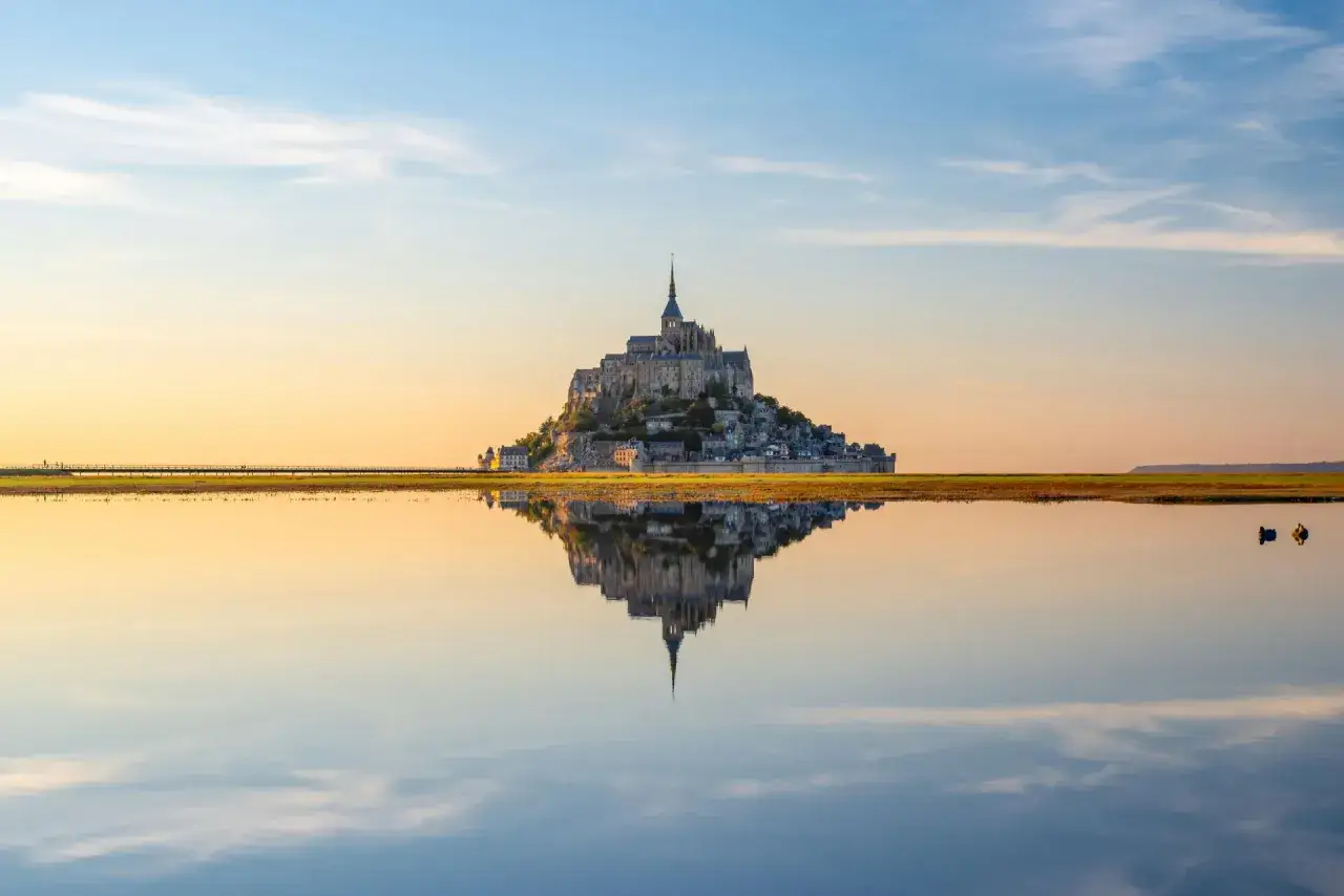 Mont Saint Michel, un'isola rocciosa con un'abbazia medievale, si specchia in uno specchio d'acqua calmo al tramonto.