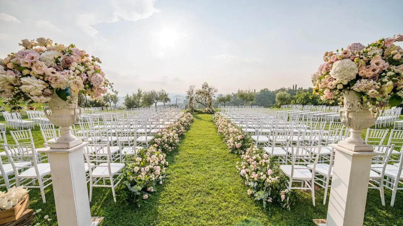 Un incantevole allestimento per un matrimonio di venerd&igrave;, con file di sedie bianche su un prato verde, decorate da fiori rosa e bianchi, sotto un cielo luminoso.