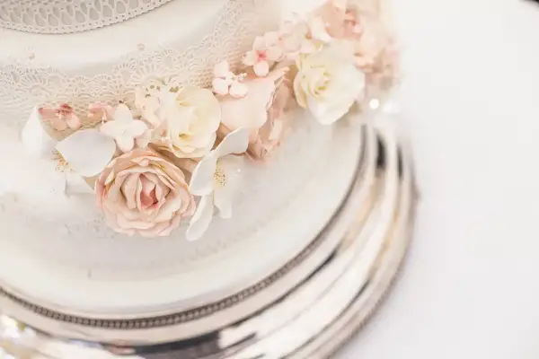 Dettaglio di una torta nuziale a più piani, decorata con pizzo e fiori di zucchero rosa e bianchi, su un elegante vassoio argentato.