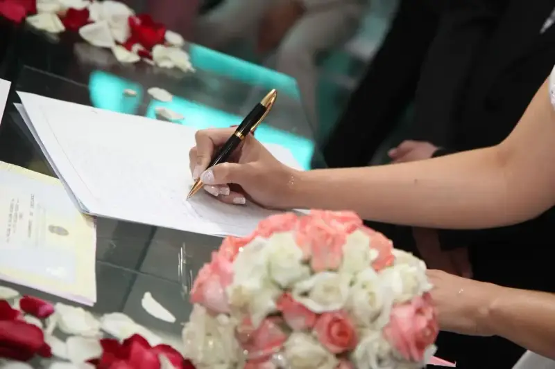 Mano di una sposa che firma un documento, forse un accordo di separazione dei beni, con un bouquet di rose in primo piano.