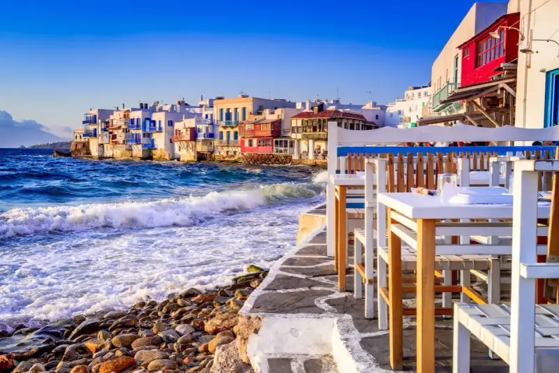 Case vacanza a Mykonos, un'idea per dove andare a vivere all'estero con pochi soldi. Case bianche sul mare, onde che si infrangono sugli scogli, tavoli di un ristorante.